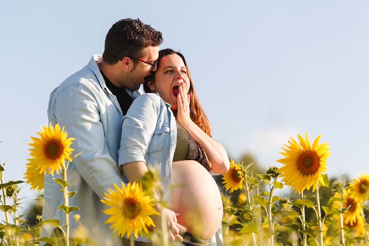 sesión de embarazo en exteriores, foto divertida mujer embarazada con barriga al aire, junto al marido dentro de un campo de girasoles