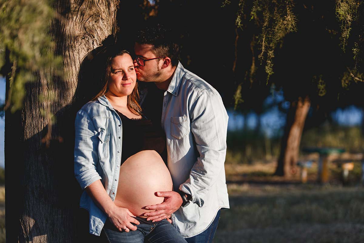 reportaje de fotos de embarazo en pareja, mujer embarazada junto a un árbol, con su pareja besándola en la mejilla y con la mano en su barriga